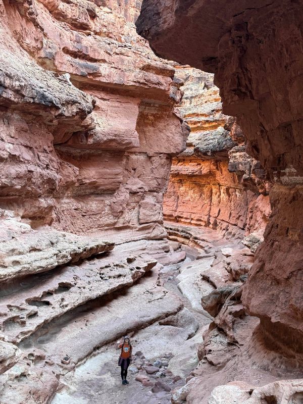 Solo hiker standing in the middle of a narrow, winding slot canyon with red rock walls and layered textures. This image showcases a striking destination along a Glen Canyon hiking route.