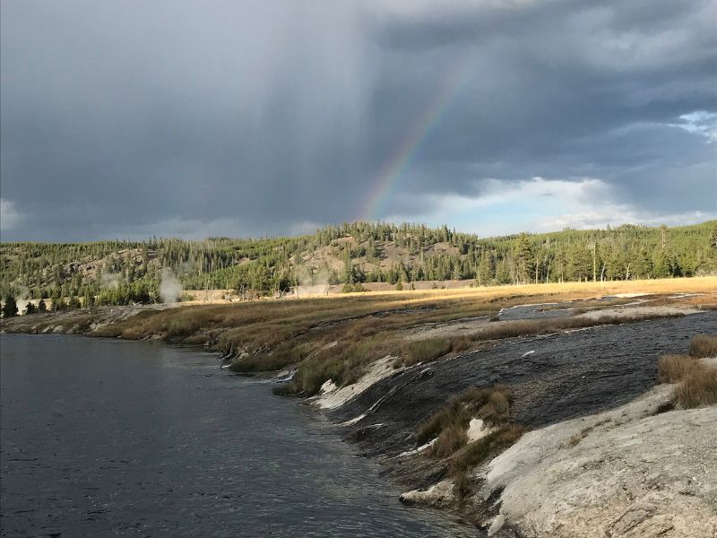 A faint rainbow appears beneath dark storm clouds over a Yellowstone river valley with a small geothermal feature steaming in the distance.