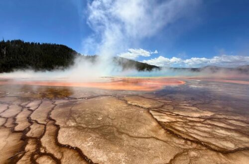 Ground level view across Grand Prismatic Spring with orange and brown microbial mats leading toward turquoise water and drifting steam.