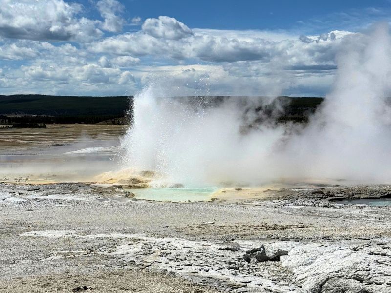 Yellowstone geyser erupts beside a steaming geothermal pool under a blue sky filled with large white clouds.