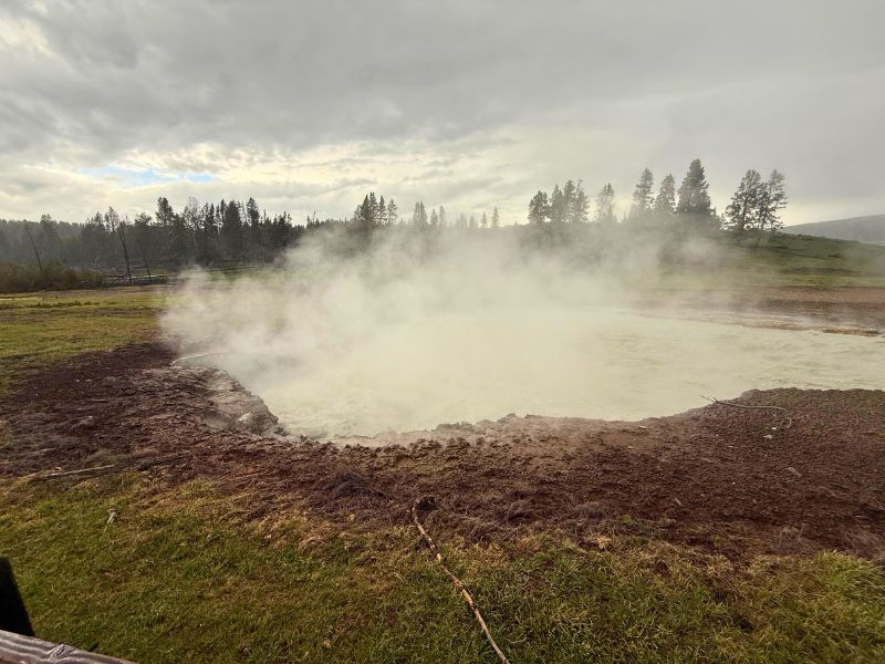 Steam rises from a pale geothermal pool in Yellowstone while dark storm clouds hang over the surrounding meadow and pine forest.