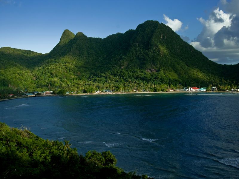 Towering, steep green mountain peaks rise above a deep blue coastal bay in American Samoa. A narrow strip of shoreline with palm trees and small buildings sits at the base of the densely forested mountains.