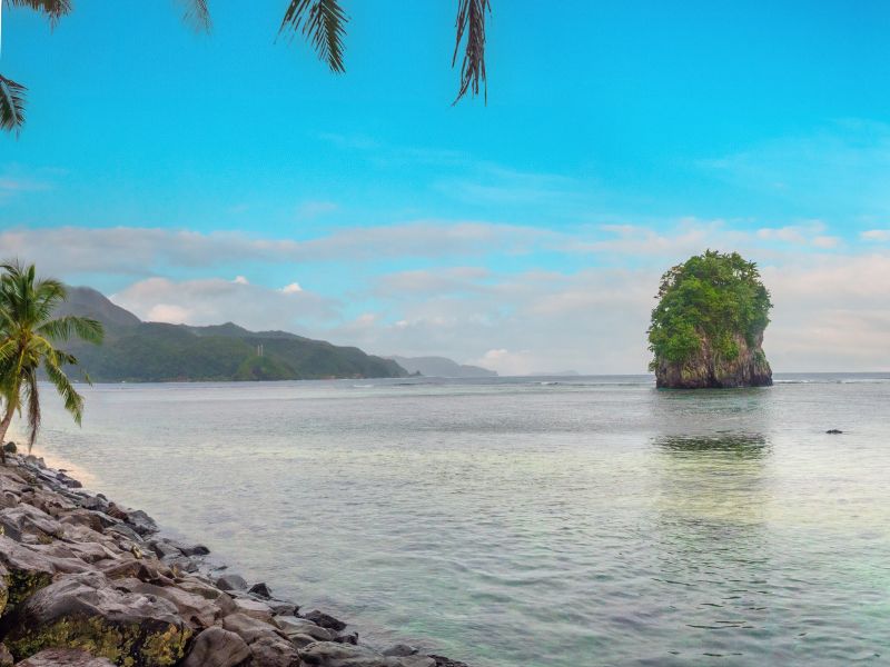 A solitary tree covered rocky islet stands in calm ocean water near the National Park of American Samoa. A rocky shoreline lined with palm trees fills the foreground, while distant green mountains stretch along the coast beneath a bright blue sky.