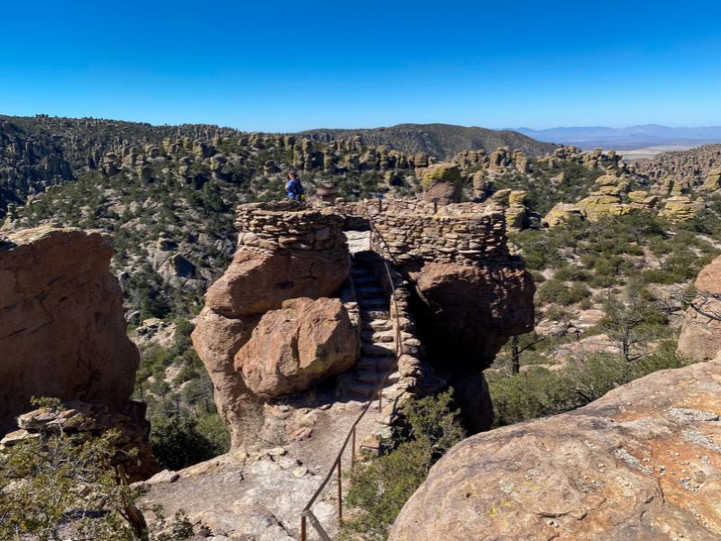 A narrow stone staircase with metal railings leads to a rocky overlook at Chiricahua National Park in Arizona. A visitor stands on the elevated platform overlooking a vast landscape of towering rock pinnacles and desert mountains beneath a clear blue sky.