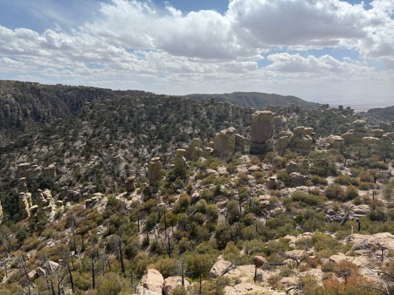 Wide scenic overlook of Chiricahua National Monument showing a rugged landscape filled with tall stone pinnacles, rounded rock formations, and scattered green trees. Layered hills extend across the horizon beneath a sky filled with large white clouds.