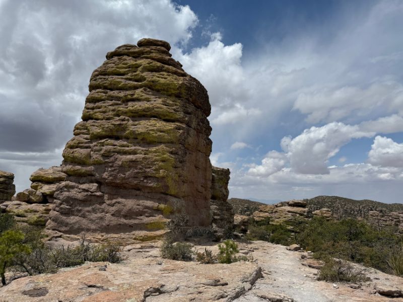 A massive freestanding rock tower with rounded layers of weathered stone dominates the foreground at Chiricahua National Monument. Patches of green lichen cover the rock surface, while smaller formations and distant rocky hills appear beneath a dramatic cloudy sky.