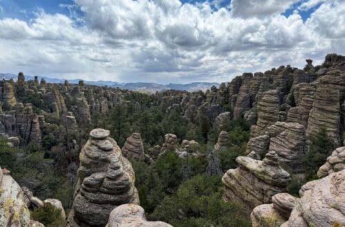 Panoramic view across a forested canyon filled with hundreds of unusual stacked rock pinnacles and rounded stone towers at Chiricahua National Monument. Large layered formations frame the foreground, while dense green trees fill the valley and distant mountains stretch beneath a partly cloudy sky.