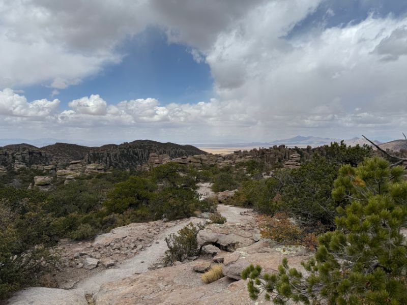 A rocky hiking trail winds downhill through shrubs and small pine trees toward an expansive landscape of weathered rock formations at Chiricahua National Monument. Distant desert mountains and a broad valley appear beneath dramatic white and gray clouds.