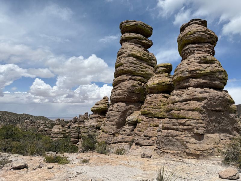 Three massive columns of layered volcanic rock rise above a rocky landscape at Chiricahua National Monument, with rounded boulders balanced near their tops. Smaller stone pillars and scattered desert vegetation surround the formations beneath a partly cloudy sky.