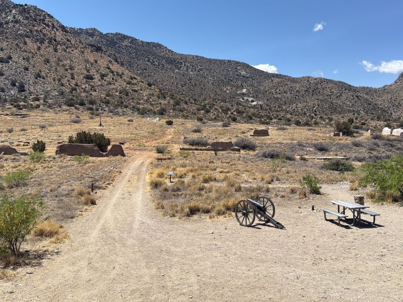 A dirt trail winds through the desert landscape of Fort Bowie National Historic Site in Arizona. Historic adobe ruins, an old cannon carriage, and a picnic table sit among dry grasses and shrubs with rugged mountains in the background.