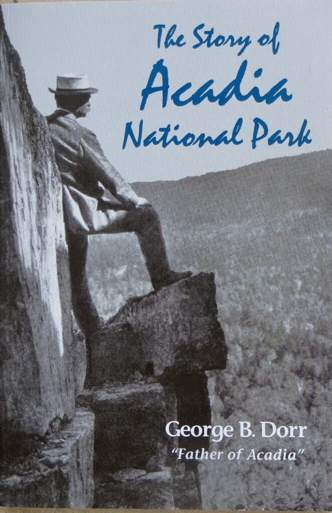 Book cover of "The Story of Acadia National Park" by George B. Dorr, known as the "Father of Acadia." A black and white historical photograph shows a man in a hat and suit standing on rocky terrain overlooking a mountainous landscape, with the title in blue lettering.